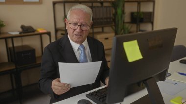 Takım elbiseli kıdemli adam, bilgisayar, notlar ve bitkilerle çevrili ofis ortamında profesyonel bir iş ortamını yansıtan belgeler okuyor..