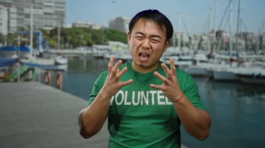 Yeşil gönüllü gömlekli genç adam, güneşli bir günde deniz kenarındaki limanda arka planda görünen yatlarla duygularını ifade ediyor..