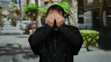 Çapraz kolyeli siyah cüppeli Asyalı erkek rahip sokak ortasında dikiliyor yüzünü örtüyor ve gülümsüyor..