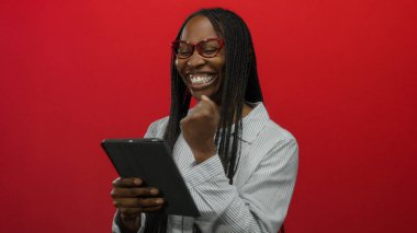 Örgülü ve gözlüklü Afrikalı kadın gülümsüyor ve elinde tabletle parlak kırmızı arka planda teknoloji zevkini ve modern yaşam tarzını gösteriyor..