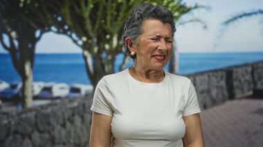 Gri saçlı yaşlı kadın, deniz kenarında ağaçlar ve okyanus manzaralı bir yerde hoşnutsuz bir yüz yapıyor..