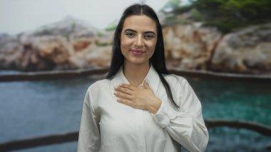Deniz kenarında kalbi üzerinde duran bir kadın, kayalık kayalıklara ve sakin bir okyanusa karşı kendine güveni yayıyor, açık havayı kucaklıyor..