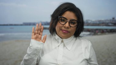 Sahilde gözlüklü genç bir kadın plaj arka planında beyaz bir gömlek giyiyor, çeşitliliği ve vücut pozitifliğini temsil ediyor..