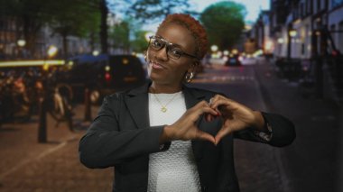 Gün batımında, parıldayan trafik ışıkları arasında, canlı şehir caddesinde elleri olan, gözlüklü gülümseyen kadın kalp şeklini alır..