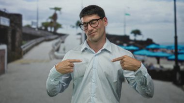 Beyaz gömlekli ve gözlüklü bir adam göğüs göğse işaret ediyor ve kıyı şeridinde sokakta gülümsüyor. Kendine güveniyor..