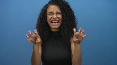 Mavi stüdyoda pençe jesti ve gülümseyen yüzüyle gözlük takan bir kadın; neşe dolu bir eğlence.