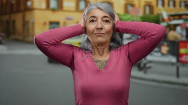 Gri saçlı bir kadın, sokakta pembe bir kazak giyiyor. Düşünceli bir ifade takınıyor. Şehir içi stresi ya da bir şehirde tefekkür izlenimi veriyor..