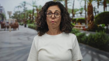 Orta yaşlı kıvırcık saçlı ve gözlüklü İspanyol kadın sokakta neşeli bir sırıtışla ağzını açar..