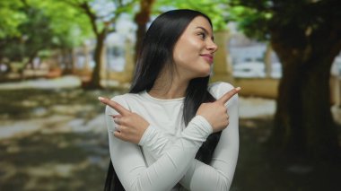 Açık yeşil ağaçlarla çevrili güneşli bir parkta gülümserken, beyaz tişörtlü, uzun siyah saçlı bir kadın parmaklarını zıt yönlere çeviriyor.