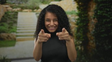 Genç kadın, gözlük takmış parmaklarıyla gözlere çerçeve çiziyor ve sokak basamaklarında kamerayı işaret ediyor; oyuncu merak.