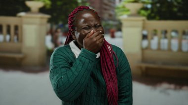 Şehir caddesinde ağzı kapalı bir şekilde gülen bir kadın yeşil bir kazak ve pembe örgülü saçlarla şehir ortamını ve güneş ışığını gösteren bulanık bir arka planı var..