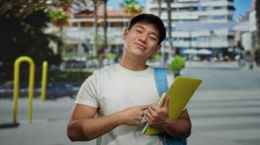 Sırt çantası ve klasörleri olan genç Çinli adam güneşli bir şehir caddesinde olumlu ve gençliği yansıtan bir şehir ortamında başparmak kaldırıyor..