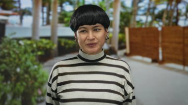 Çizgili kazaklı genç bir kadın açık havada gülümsüyor. Elleri açık, sakin bir bahçe ortamında güvenle poz veriyor..