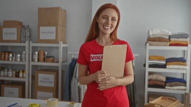 Bağış kutuları, giysiler ve malzemelerle çevrili hayır kurumunda elinde pano tutan gülümseyen kızıl saçlı kadın, bir bağışta bulunma merkezindeki gönüllü işleri simgeliyor..