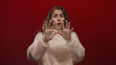 Kadın, canlı kırmızı bir arkaplan karşısında şaşırmış, rahat bir süveter giyiyor, duygularını ifade eden gözler ve ellerle yakalıyor, yaratıcı projeler için tamamen izole edilmiş..