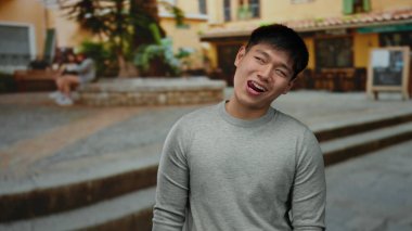 Şehrin arka planında neşeli bir şekilde gülümseyen genç bir adam, şehir manzarasını günlük atmosferiyle, güneşli günüyle ve rahat ifadesiyle yakalıyor..