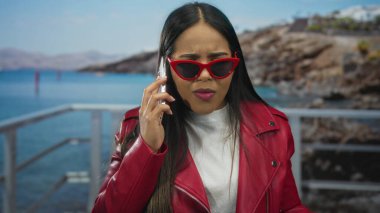 Telefon kullanan bir kadın, deniz kenarında elinde akıllı bir telefonla gezen Latin kökenli bir gencin bulunduğu bir plaj zeminine kızgın bir bakış atıyor..