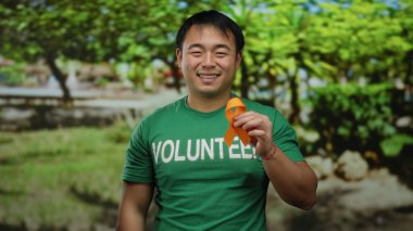 Yeşil gönüllü tişörtü giyen genç adam arka planda yemyeşil ağaçlarla dolu bir parkta beklerken elinde turuncu bir kurdele tutuyor..