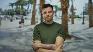 Şehir sokağında palmiye ağaçlarıyla dikilen, yeşil tişörtlü, kolunda dövme olan, düşünceli ve kendini kucaklayan bir adam..