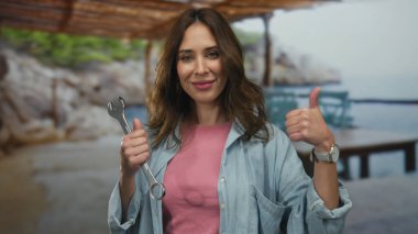 İspanyol kadın bir İngiliz anahtarı tutuyor, baş parmağıyla gülümsüyor, arka planda deniz olan bir sahilde duruyor, işi ve rahatlamayı harmanlıyor..
