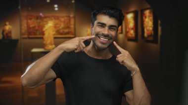 Sanat galerisinde sıcak ışıklandırma ve sergilenen heykeller ve tabloların ortasındaki yanakları işaret eden adam; mutluluk.