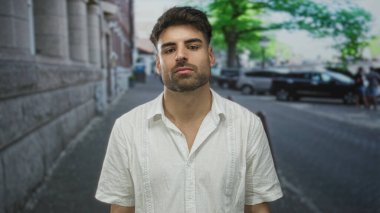 Sakallı bir adam taştan duvarın yanındaki şehir sokağında duruyor ve güneş ışığı altında park etmiş bir araba yumuşak gölgeler bırakıyor..