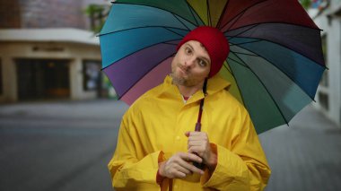 Sarı yağmurluklu adam şehir caddesinde renkli bir şemsiye tutuyor.