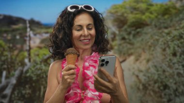 Parkta bikini ve çiçek giymiş İspanyol bir kadın güneşli bir günde selfie çekerken dondurmanın tadını çıkarıyor..