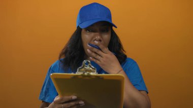 Mavi şapkalı kurye kadın stüdyoda elinde bir pano ve ağza takılı bir kalem tutuyor..