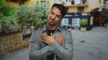 Genç bir adam, İspanyol asıllı, açık havada gülümsüyor, kolları çaprazlanmış bir şekilde şehir merkezinde binaların arka planında, mutluluğu ve rahatlığı aktarıyor..