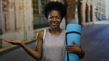 Şehir caddesinde elinde mavi bir yoga minderiyle duran bir kadın, arka planda modern kentsel mimari ile açık hava antrenmanına hazır olduğunu ve özgüvenini gösteriyor..