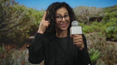 Orta yaşlı İspanyol kadın parkta mikrofonu tutarken parmaklarıyla kafasına dokunuyor..