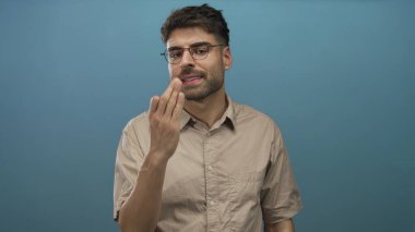 Mavi stüdyoda gözlüklü, büzüşmüş dudaklı ve çimdiklenmiş parmaklı İspanyol bir adam jest yapıyor..