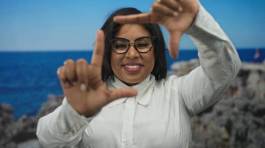 Kumsalda gülümseyen bir kadın, deniz kenarında bir zemine karşı elleriyle bir çerçeve oluşturuyor, gözlük takıyor ve beyaz bir gömlek giyiyor, açık deniz kenarında güneşli bir günün tadını çıkarıyor..