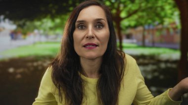 Sarı kazaklı kadın, güneşli park ortamında, yemyeşil ile çevrili, meraklı ve ince yüz hareketleriyle meşgul..