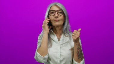 Gri saçlı yaşlı kadın akıllı telefondan konuşuyor ve pembe arka planda baş parmağıyla hareketler yapıyor, gözlük takıyor ve kolunda dövmeleri olan beyaz bir gömlek giyiyor..