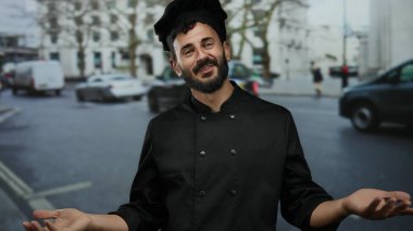 Siyah üniformalı İspanyol şef, kalabalık bir caddede durup, yoldan geçenlere gülerek enerjik bir şekilde el hareketi yapar..