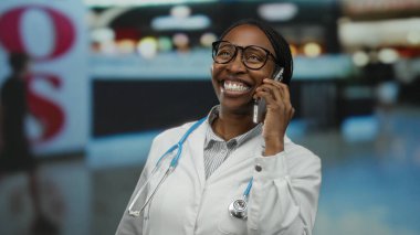 Beyaz önlüklü, steteskopla gülümseyen bir kadın kapalı hava alanında akıllı telefondan konuşuyor. Profesyonel iletişim ve seyahat temasını öneriyor..