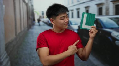 Gündüz vakti şehir caddesinde plaka taşıyan genç adam, kendine güvenerek gülümsüyor, sürücü statüsünü gösteriyor, kırmızı gömlek giyiyor, arka planda şehir ortamı var..