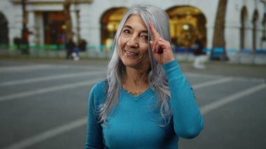Gri saçlı kadın gülümsüyor ve bir şehir caddesinde işaret ediyor, mavi giyiniyor, arka planda bulanık bir bina var..