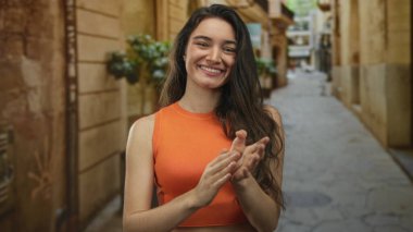 Turuncu giysili bir kadın açık gökyüzünün altında güneşli kumtaşı binasının yanında el çırpıyor..