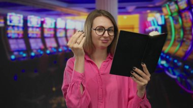 Gözlük takan, kitap okuyan, arka planda kumar makineleri olan canlı bir kumarhanede para hareketi yapan bir kadın..