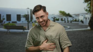 Sakallı genç adam, güneşli bir caddede bej gömlekli, şehir arka planında beyaz binalar ve arkasında park bankları olan neşeli bir şekilde gülümsüyor.