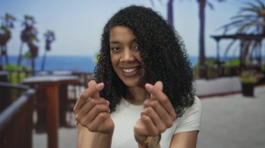 Kıvırcık saçlı, gülümseyen ve kalp hareketi yapan bir kadın parmaklarıyla sokağın ahşap parmaklıklarının yanında, sevgi dolu..