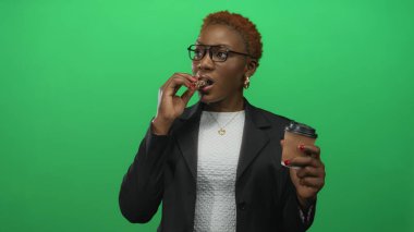 Stüdyoda granola yiyen ve hazır kahve fincanı tutan bir kadın siyah ceket ve gözlük takıyor..