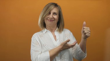 Stüdyoda elleri açık sunan ve beyaz bluz giyen turuncu arka planlı bir kadın..