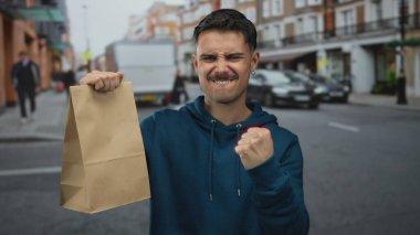 Şehir caddesinde bir kese kağıdıyla kutlama yapan genç bir adam, dışarıda mavi kapüşonlu bir kapüşonlu giyen, yumruk pompasıyla neşeyi ifade ediyor..
