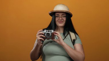 Kameralı kadın gezgin canlı turuncu arka planda poz veriyor safari şapkası takıyor güven ve macera ruhu yayıyor.