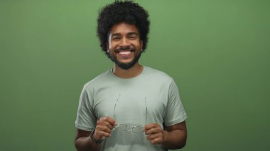Afro-Amerikan bir adam gülümserken, yeşil duvarlara karşı gözlüklerini indiriyor, mutluluk ve güven yayıyor..