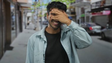 Genç İspanyol adam, parlak şehir caddesinde park etmiş arabalarla dururken gözlerini eliyle kapatıyor; utanç..
